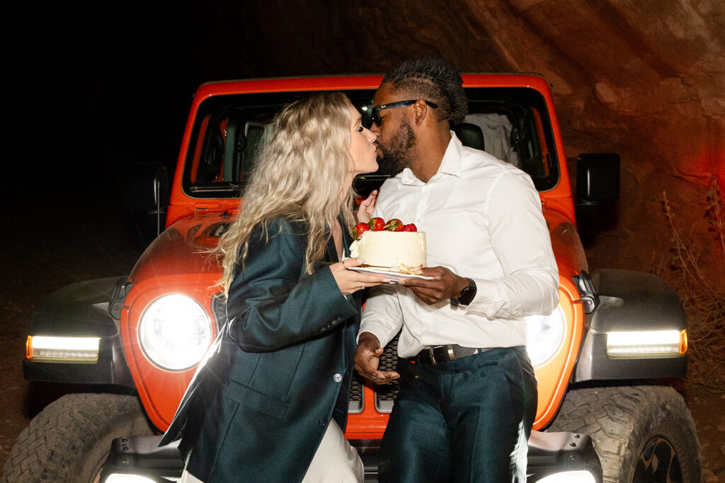 bride and groom sharing a small elopement cake sitting in front of jeep headlights