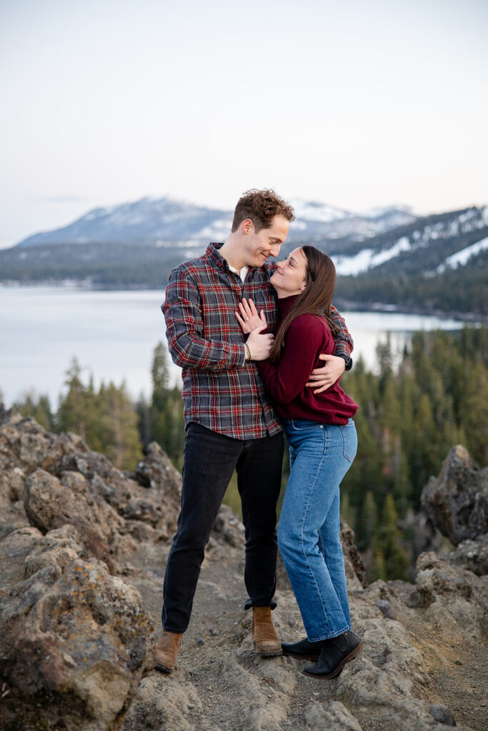 A couple shares a sweet moment  standing on a rocky cliff with a view of Lake Tahoe and snow-covered peaks behind them during golden hour Eagle Rock engagement photos