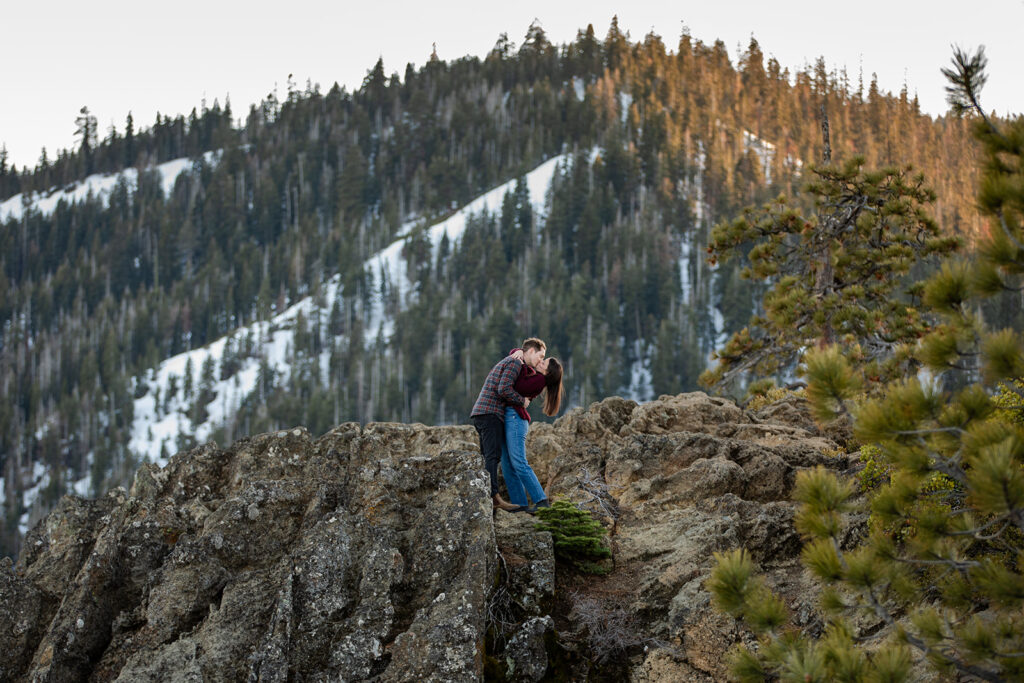 On a rugged cliff at Eagle Rock, the couple kisses passionately with a panoramic view of pine trees and steep snowy mountains in the background.