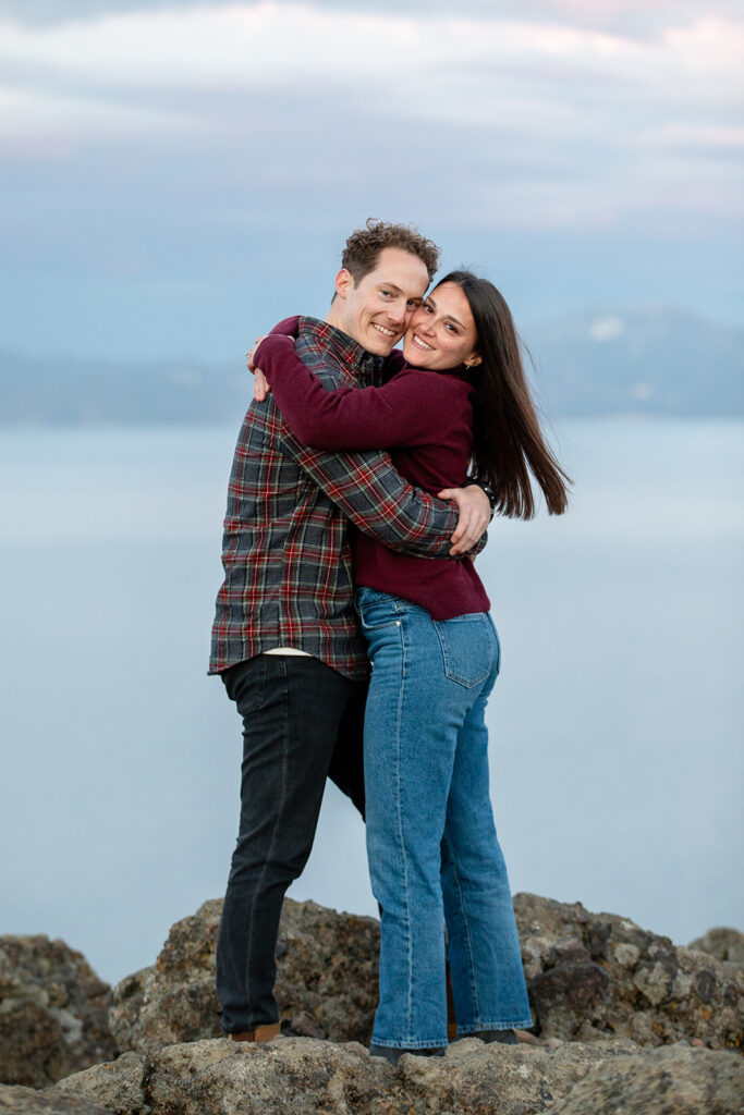 A couple shares a sweet moment  standing on a rocky cliff with a view of Lake Tahoe and snow-covered peaks behind them during golden hour Eagle Rock engagement photos