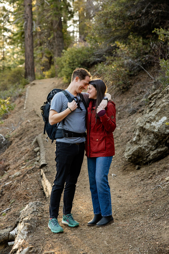 The couple stands on a wooded hiking trail, cozying up and smiling at each other at Eagle Rock