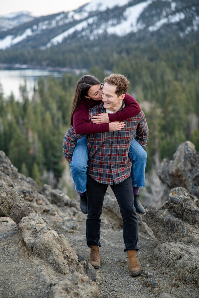 Eagle Rock engagement session with a playful piggyback ride, the couple is smiling joyfully while hiking on a rocky trail with snow-capped mountains and evergreen trees in the background near Lake Tahoe.