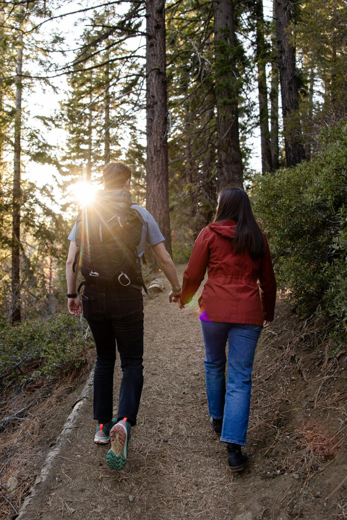 Walking hand in hand on a forest path, the couple is seen from behind as sunlight streams through the trees, casting a warm glow over the scene at Eagle Rock, Lake Tahoe