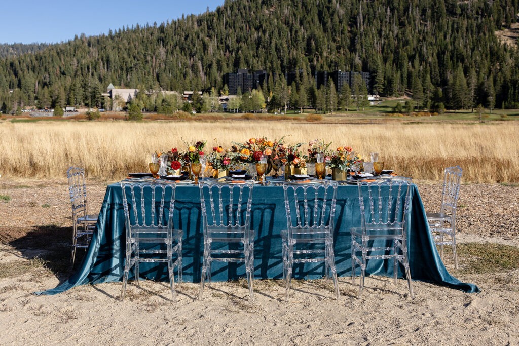 Long reception table set in a golden field with teal linen, amber and crystal glassware, and lush floral centerpieces in gold vases, surrounded by clear chairs