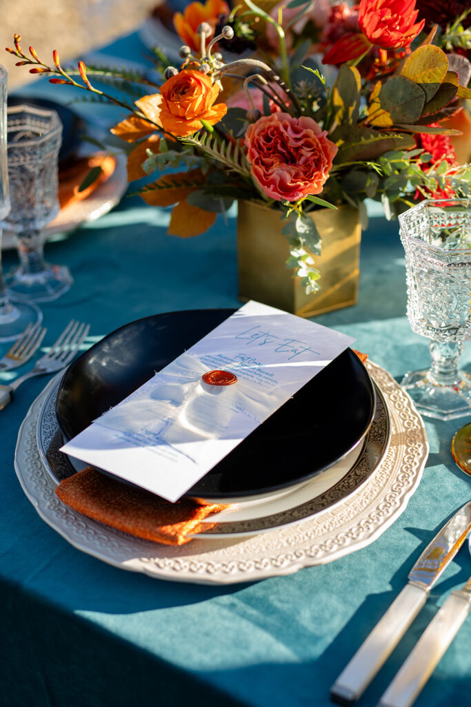 Close-up of a wedding place setting with layered plates, orange napkin, black bowl, and a menu tied with ribbon and wax seal, alongside vibrant orange and pink florals