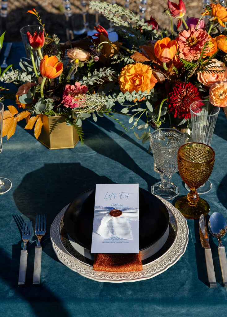 Close-up of a wedding place setting with layered plates, orange napkin, black bowl, and a menu tied with ribbon and wax seal, alongside vibrant orange and pink florals