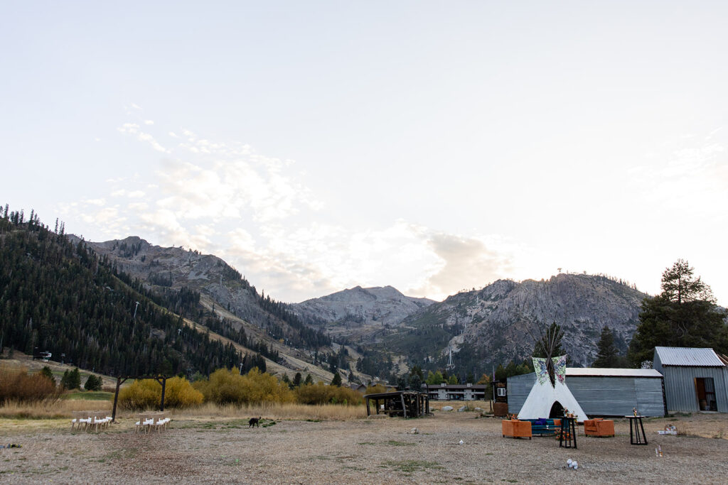 Wide landscape view of Olympic Valley with rugged mountain peaks, a tipi-style tent, and rustic structures nestled in a serene meadow at sunset at Olympic Valley Stables