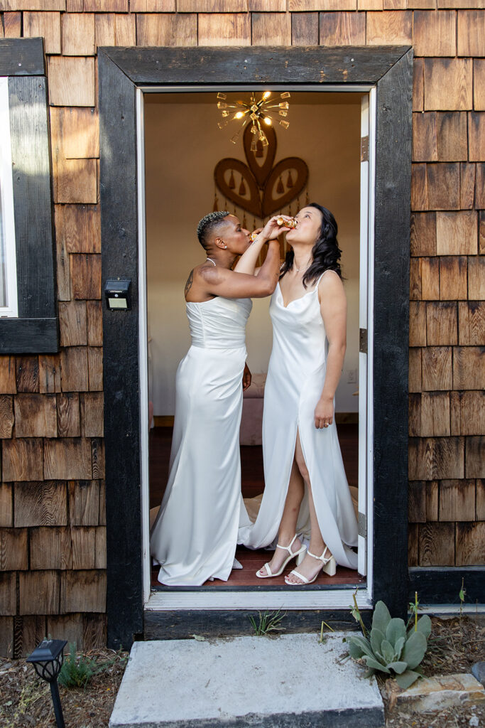 The two brides share a fun toast, arms intertwined, while standing in a doorway framed with rustic wood and mid-century modern details inside