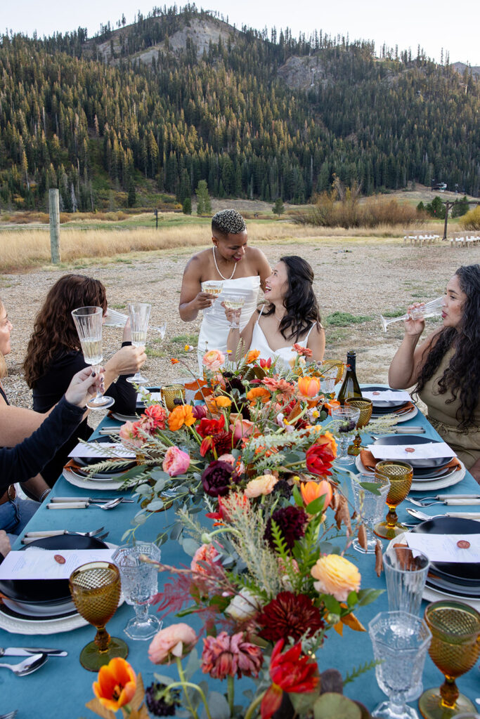 One bride stands beside her seated partner, both smiling with champagne glasses raised, surrounded by loved ones at a vibrant outdoor reception table set with bold florals and vintage glassware