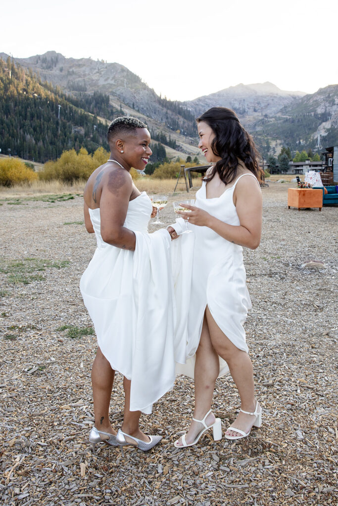 an lgbtq elopement couple hold hands while sipping champagne, standing in their wedding dresses against a backdrop of mountains and golden foliage at Olympic Valley Stables