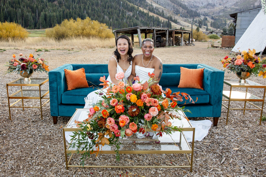 The brides sit on a teal velvet couch with orange pillows, laughing joyfully, with a lush floral arrangement bursting with autumn tones in front of them