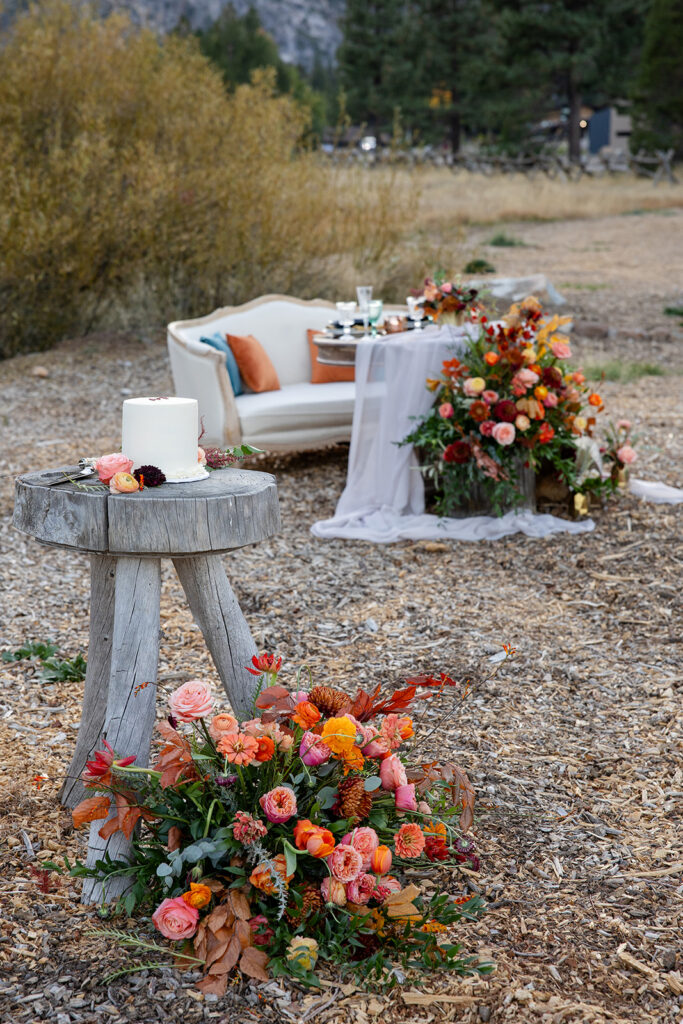 Romantic sweetheart table with an ivory settee and a cascade of orange, pink, and burgundy flowers on flowing white fabric, set in a mountain meadow at Olympic Valley Stables