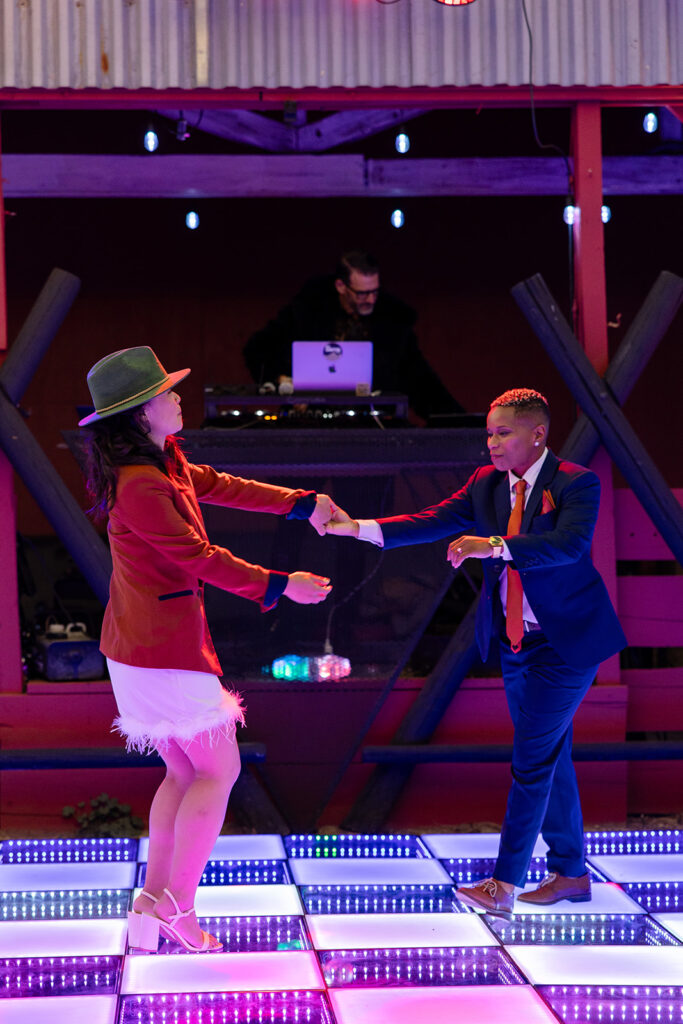 The couple dances playfully on a lit-up dance floor—one wearing a green hat and rust blazer with a feathered white mini dress, the other in a navy suit