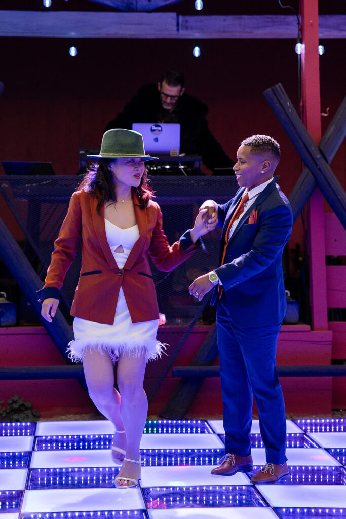 The couple dances playfully on a lit-up dance floor—one wearing a green hat and rust blazer with a feathered white mini dress, the other in a navy suit