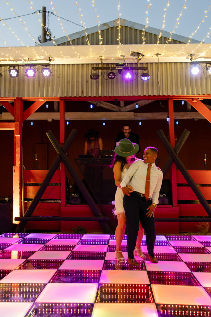 The couple dances playfully on a lit-up dance floor—one wearing a green hat and rust blazer with a feathered white mini dress, the other in a navy suit