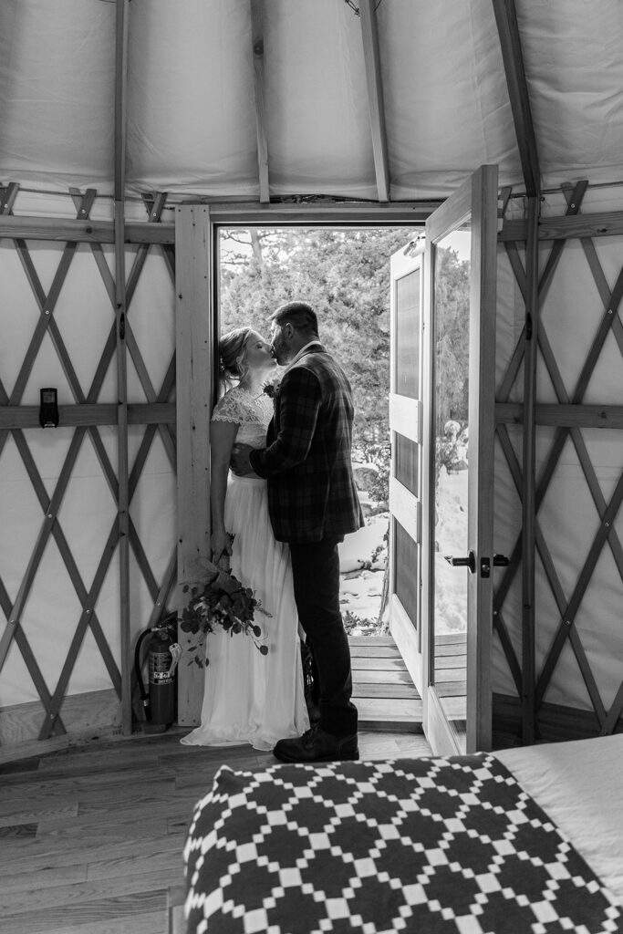 The bride and groom stand close at the yurt doorway, exchanging sweet glances as golden light spills in from outside. The bride holds a small bouquet of eucalyptus.