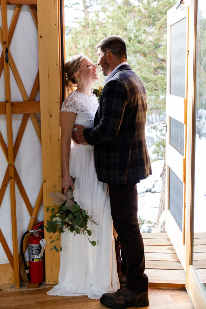 The bride and groom stand close at the yurt doorway, exchanging sweet glances as golden light spills in from outside. The bride holds a small bouquet of eucalyptus.