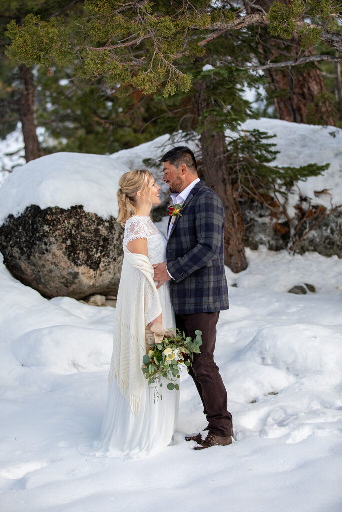 The bride and groom smile at each other while standing in deep snow among pine trees during their winter elopement in Lake Tahoe