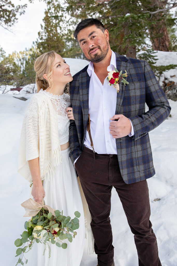 The bride and groom smile at each other while standing in deep snow among pine trees during their winter elopement in Lake Tahoe
