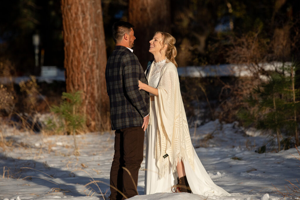 The bride and groom smile at each other while standing in deep snow among pine trees, with the bride wrapped in a shawl and holding a wintry bouquet